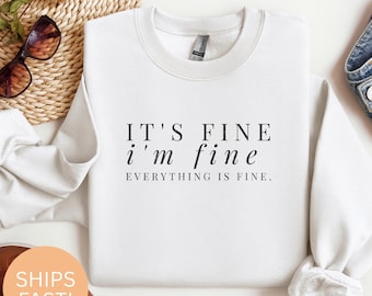 Peut inclure: Sweat-shirt blanc à col rond avec le texte "IT'S FINE i'm fine EVERYTHING IS FINE" en noir. Le sweat-shirt est plié, avec des lunettes de soleil et un panier en osier en arrière-plan. Une paire de jeans est visible à droite.
