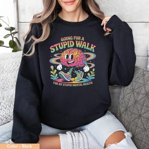 Op de afbeelding: Zwarte sweatshirt met een kleurrijke afbeelding van een lopend brein met de tekst "Going for a Stupid Walk" en "For My Stupid Mental Health". Het brein lacht en draagt sneakers. Het ontwerp heeft een retro, cartoon stijl.
