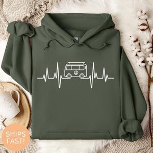 May include: An olive green hoodie featuring a white graphic of a camper van integrated into a heartbeat line. The van includes a peace symbol. The hoodie has a hood and a drawstring.