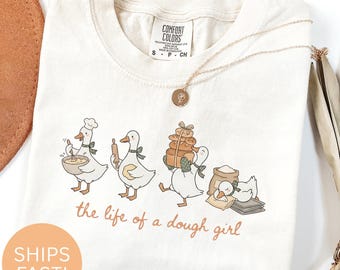 May include: Cream-colored t-shirt with a whimsical illustration of geese baking. The geese are depicted with baking tools and loaves of bread. The text "the life of a dough girl" is printed below the illustration. A gold necklace with a pendant is also visible.