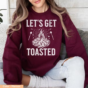 Op de afbeelding: Bordeaux sweatshirt met de witte tekst "LET'S GET TOASTED". De afbeelding toont een kampvuur met vlammen, marshmallows op stokjes en sterren. Een casual top, perfect voor kampeerliefhebbers.