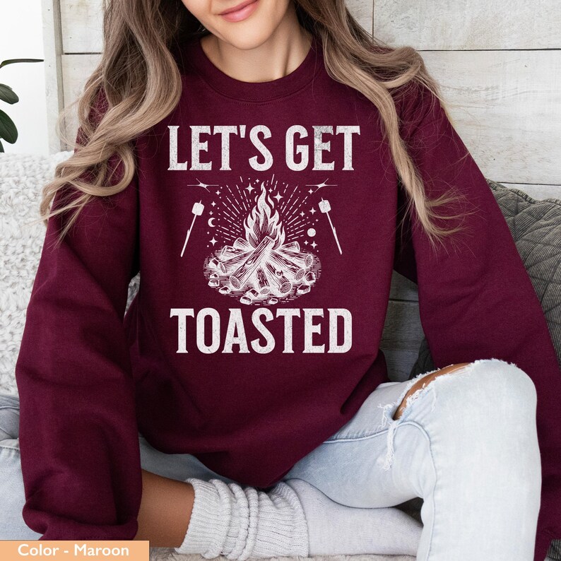 Op de afbeelding: Bordeaux sweatshirt met de witte tekst "LET'S GET TOASTED". De afbeelding toont een kampvuur met vlammen, marshmallows op stokjes en sterren. Een casual top, perfect voor kampeerliefhebbers.