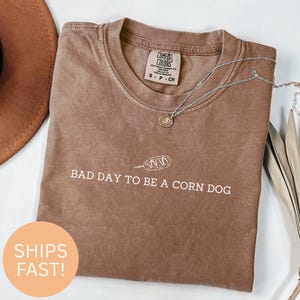 Op de afbeelding: Bruin Comfort Colors T-shirt met de witte tekst "BAD DAY TO BE A CORN DOG". Een zilveren ketting met een kleine hanger ligt op het shirt. Het shirt heeft een klein label met de tekst "COMFORT COLORS".