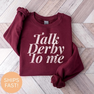 May include: A maroon sweatshirt with the phrase "Talk Derby To Me" in white, distressed lettering. The sweatshirt is displayed on a light wood-grain surface. The sleeves are folded. The sweatshirt is a Gildan brand.