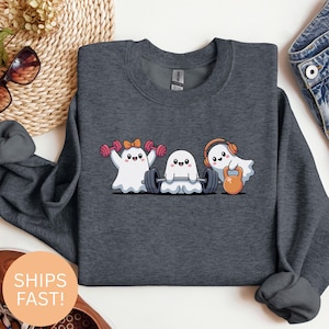 Könnte beinhalten: Dunkelgraues Sweatshirt mit einem Cartoon-Design von drei trainierenden Geistern. Ein Geist hebt Kurzhanteln, ein anderer eine Langhantel und der dritte eine Kettlebell. Die Geister haben lächelnde Gesichter und tragen Accessoires.