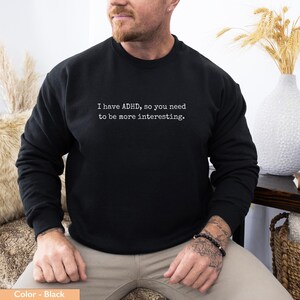 May include: Black crew neck sweatshirt with the white text: "I have ADHD, so you need to be more interesting." The sweatshirt is worn by a person. The person is wearing a bracelet on their wrist. The background includes a vase with decorative wheat.