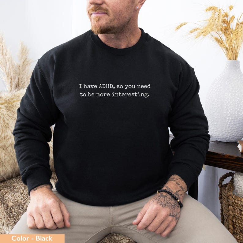 May include: Black crew neck sweatshirt with the white text: "I have ADHD, so you need to be more interesting." The sweatshirt is worn by a person. The person is wearing a bracelet on their wrist. The background includes a vase with decorative wheat.