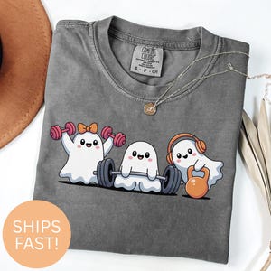 May include: Gray Comfort Colors t-shirt featuring three cartoon ghosts lifting weights. One ghost lifts pink dumbbells, another lifts a barbell, and the third lifts an orange kettlebell. The shirt has the text "Comfort Colors" and "S - P - CH" printed on the tag.