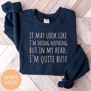 Può includere: Felpa blu navy con testo bianco: "IT MAY LOOK LIKE I'M DOING NOTHING BUT IN MY HEAD, I'M QUITE BUSY." La felpa ha girocollo e maniche lunghe. Un cerchio color pesca dice "SHIPS FAST!"