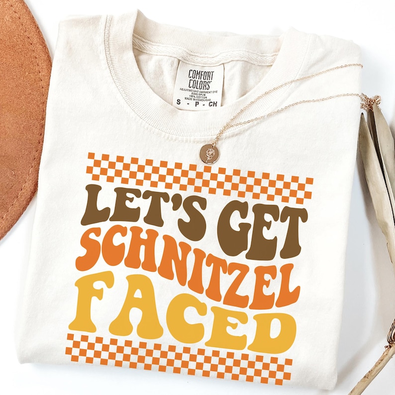 May include: Off-white t-shirt featuring the text "LET'S GET SCHNITZEL FACED" in brown, orange, and yellow, with a checkered pattern. A gold necklace with a pendant is draped across the shirt.
