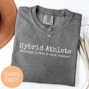 Może przedstawiać: Szara koszulka Comfort Colors z napisem "Hybrid Athlete" i frazą "(Average lifter & shit runner)". Złoty naszyjnik z wisiorkiem spoczywa na koszulce. Okrągła naklejka z napisem "SHIPS FAST!" znajduje się po lewej stronie u dołu.