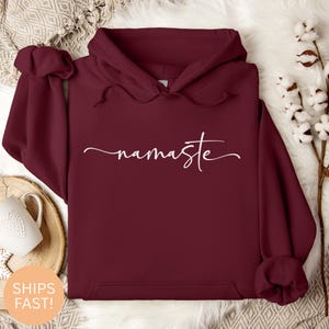 Puede incluir: Una sudadera con capucha de color granate con la palabra "namaste" en escritura blanca en el pecho. La sudadera tiene una capucha con cordón y un bolsillo delantero. El fondo incluye una taza blanca y elementos decorativos.
