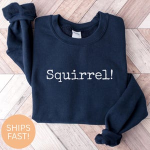 Pode incluir: Uma camisola azul marinho com a palavra "Squirrel!" impressa em branco na frente.