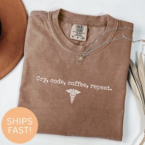May include: A brown Comfort Colors t-shirt with the text "Cry, code, coffee, repeat" and a medical symbol. The shirt has a crew neck and short sleeves. A silver necklace with a pendant is draped across the shirt.