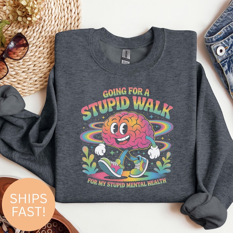 Op de afbeelding: Donkergrijze sweatshirt met een kleurrijke afbeelding van een hersen die loopt met armen en benen. De tekst luidt "Going for a Stupid Walk for my Stupid Mental Health" in een regenbooglettertype. De hersen lacht en draagt sneakers.