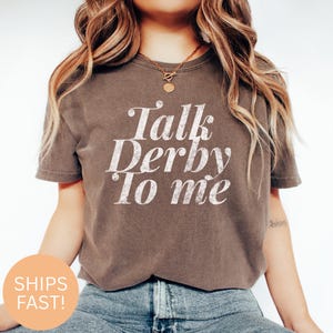 Może przedstawiać: Brązowa koszulka z napisem "Talk Derby To Me" w postarzanym białym kroju pisma. Koszulka ma okrągły dekolt i wydaje się być wykonana z miękkiego materiału. Modelka ma na sobie niebieskie dżinsy.
