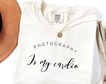 May include: A beige t-shirt with the text "Photography is my cardio" printed in black. The shirt is folded and laid flat on a white surface.