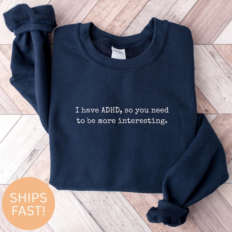 May include: Navy blue sweatshirt with the text "I have ADHD, so you need to be more interesting." in white. The sweatshirt is folded on a light brown wooden surface.