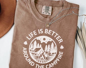 May include: A brown t-shirt with a white graphic design that says "Life is better around the campfire". The graphic features a campfire, a tent, and trees.