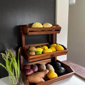 May include: A three-tiered wooden produce stand filled with fresh fruits and vegetables. The top shelf holds yellow melons, the middle shelf has lemons, limes, and kiwis, and the bottom shelf displays potatoes, sweet potatoes, and avocados. A glass of asparagus is on the left.