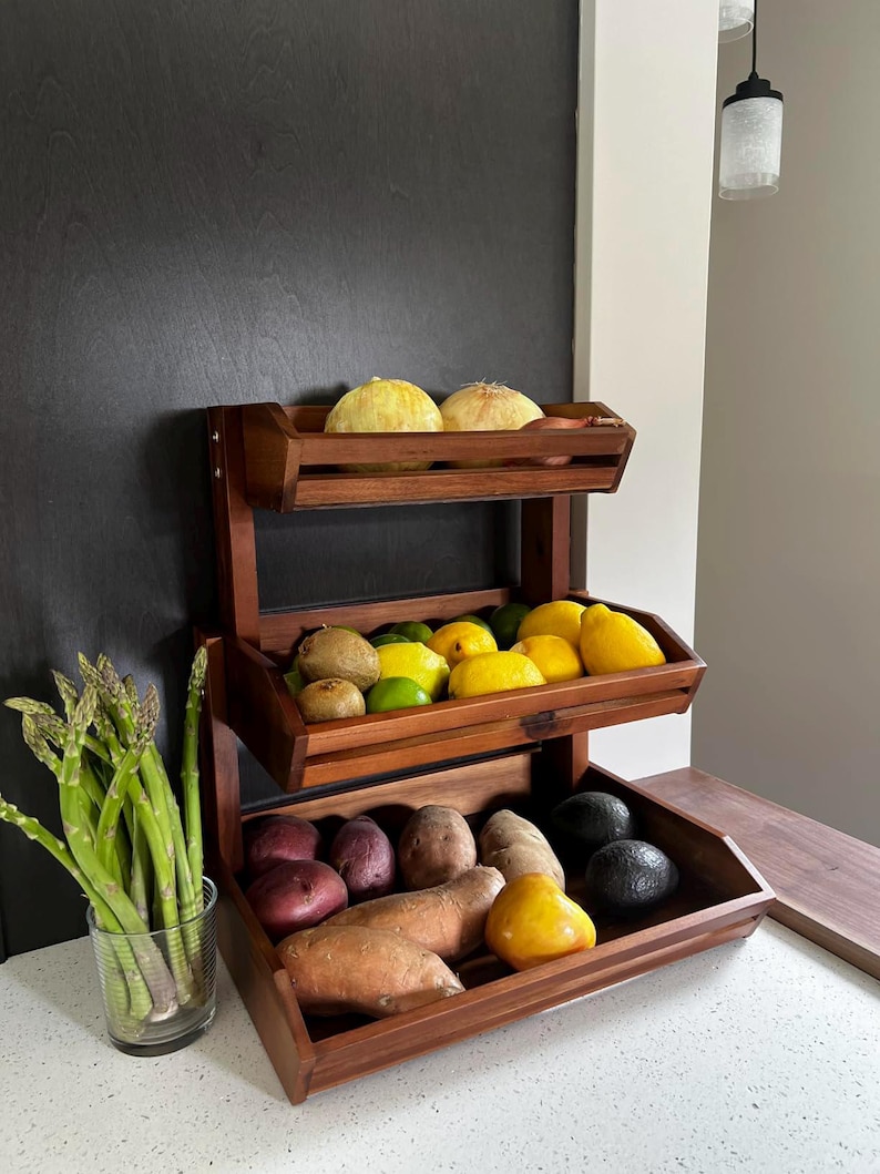 May include: A three-tiered wooden produce stand filled with fresh fruits and vegetables. The top shelf holds yellow melons, the middle shelf has lemons, limes, and kiwis, and the bottom shelf displays potatoes, sweet potatoes, and avocados. A glass of asparagus is on the left.