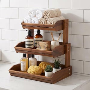 May include: A three-tiered wooden bathroom shelf unit. The top shelf holds rolled white and beige towels. The middle shelf has shampoo and conditioner bottles, soap, and toothbrushes. The bottom shelf displays body wash, lotion, a sponge, and small potted plants.