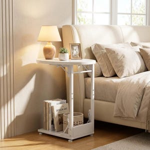 May include: A white C-shaped side table with a metal frame and two tiers. The top surface holds a beige lamp, a small potted succulent, and a framed picture. The bottom shelf contains several books and a woven basket. The table has adjustable legs.