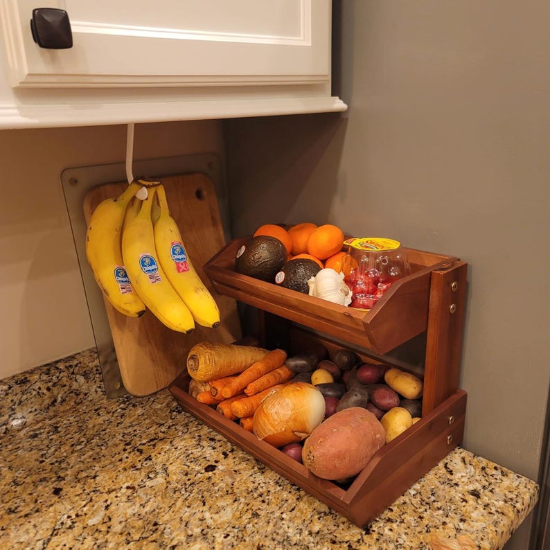May include: A two-tiered wooden produce stand filled with fresh fruits and vegetables. The top shelf holds avocados, oranges, garlic, and tomatoes. The bottom shelf contains carrots, potatoes, and an onion. Bananas hang from a hook above.
