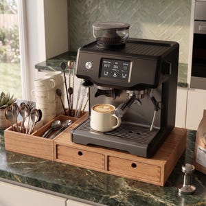 May include: A matte black espresso machine with a digital display showing 93 degrees Celsius and 25 seconds, a bean hopper on top, and a portafilter. It sits on a wooden organizer with drawers, next to a bag of Heritage Roast coffee.