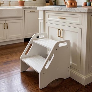 May include: A white wooden step stool with two steps. The step stool has a handle on each side and is designed to help people reach higher places. The step stool is placed on a dark wood floor in a kitchen setting.