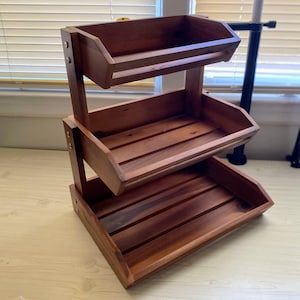 May include: A three-tiered wooden storage shelf with a rich, dark brown finish. Each tier is a tray with slatted bottoms and angled sides. The shelf is held together with metal hardware and is designed for displaying or organizing items.