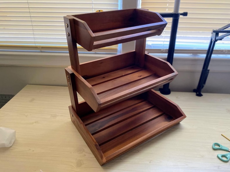 May include: A three-tiered wooden storage shelf with a rich, dark brown finish. Each tier is a tray with slatted bottoms and angled sides. The shelf is held together with metal hardware and is designed for displaying or organizing items.