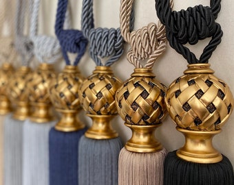 May include: Five decorative curtain tiebacks with gold finials and tassels. The tiebacks are made of braided rope in various colors, including beige, gray, blue, and black.