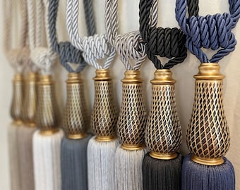 May include: Set of six decorative tassel tiebacks in various colors, including beige, gray, silver, black, and blue. Each tassel features a gold-colored metal finial with a geometric pattern.