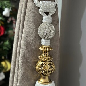 May include: White tassel tieback with a gold decorative finial and a crystal ball accent. The tieback is made of a thick, braided rope.