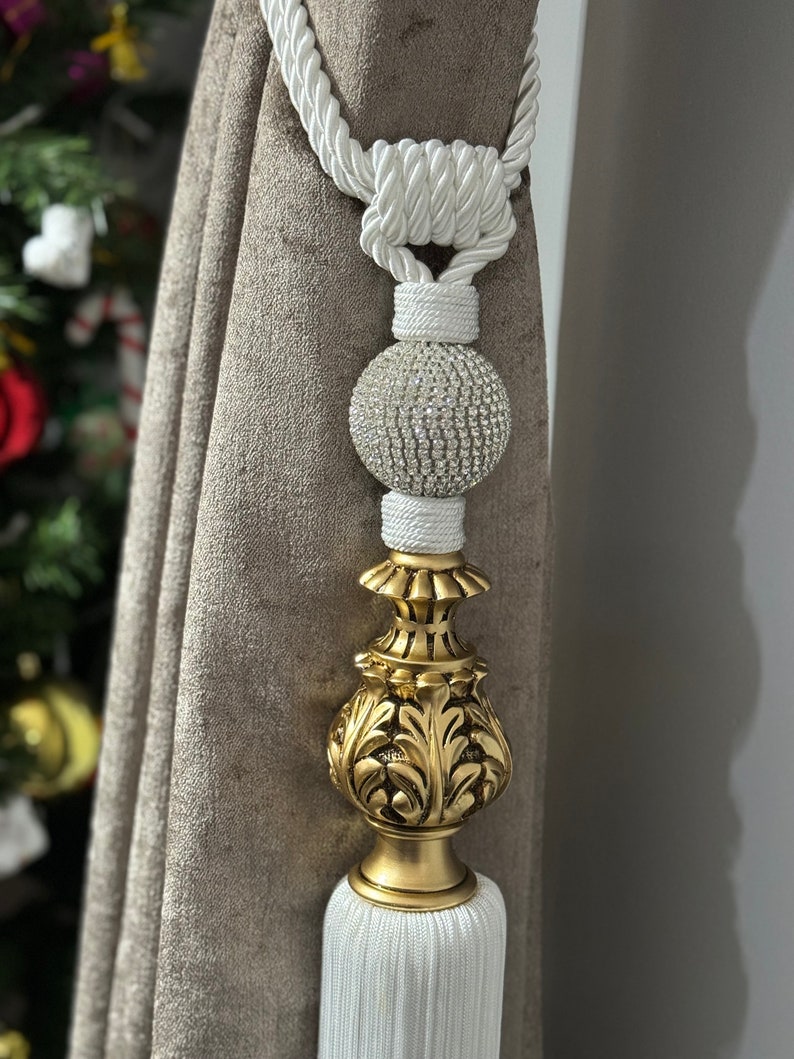 May include: White tassel tieback with a gold decorative finial and a crystal ball accent. The tieback is made of a thick, braided rope.