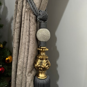 May include: Gray and gold curtain tieback with a crystal ball finial. The tieback is made of a thick, braided rope and has a tassel at the bottom.