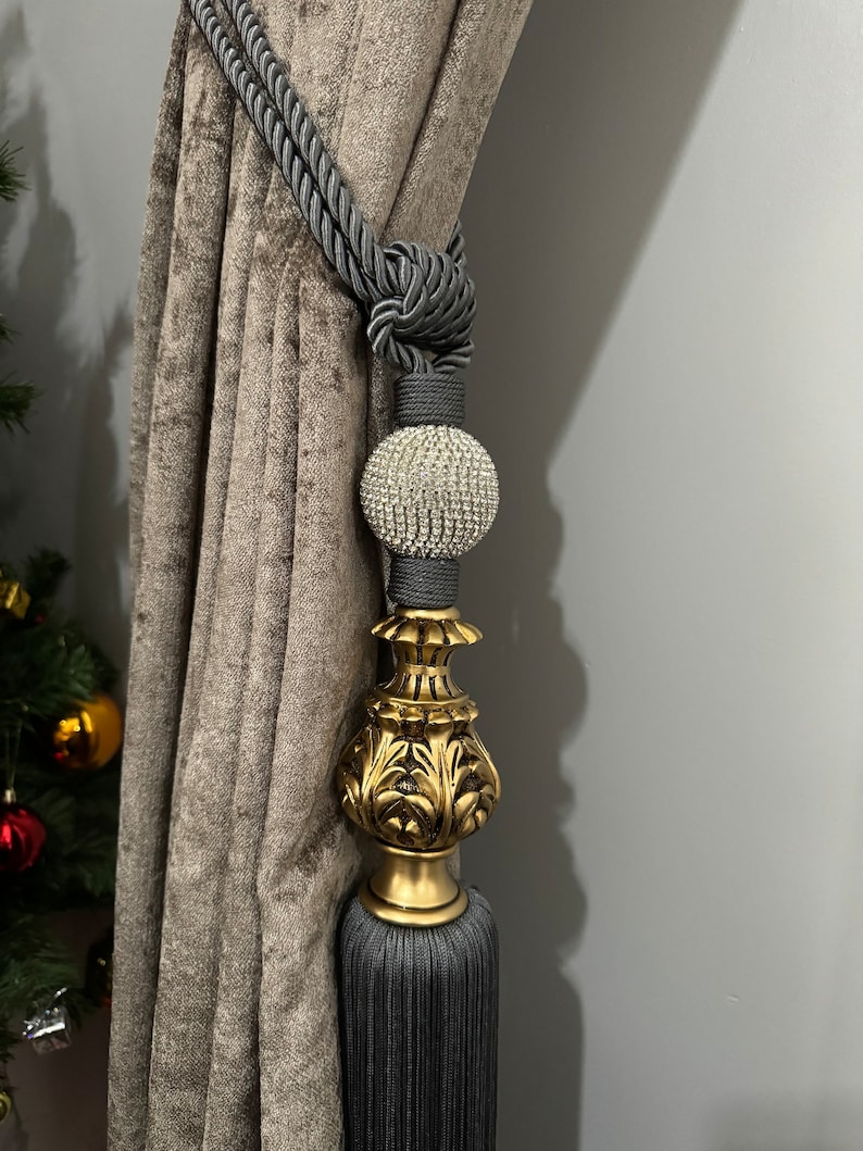 May include: Gray and gold curtain tieback with a crystal ball finial. The tieback is made of a thick, braided rope and has a tassel at the bottom.