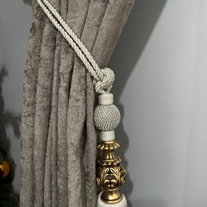 May include: A white rope curtain tieback with a gold and white tassel. The tassel has a decorative, ornate, gold top and a white, beaded ball.
