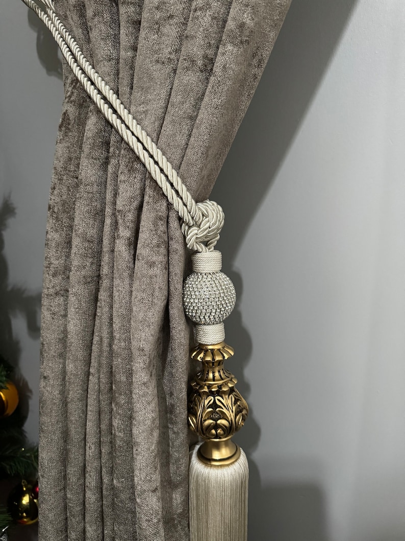 May include: A white rope curtain tieback with a gold and white tassel. The tassel has a decorative, ornate, gold top and a white, beaded ball.