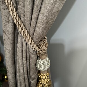 May include: A brown velvet curtain with a gold and crystal curtain tieback. The tieback features a decorative tassel and a large crystal ball.