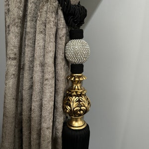 May include: Black and gold curtain tieback with a crystal ball finial. The tieback is made of a thick, braided rope and has a tassel at the bottom.