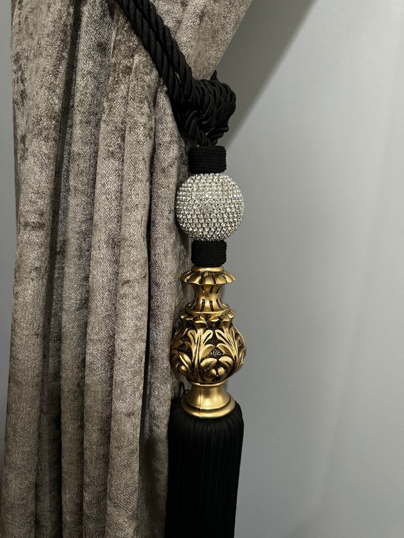 May include: Black and gold curtain tieback with a crystal ball finial. The tieback is made of a thick, braided rope and has a tassel at the bottom.