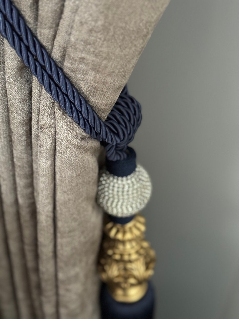 May include: A navy blue curtain tieback with a decorative tassel. The tassel features a silver bead and a gold-colored ornament.