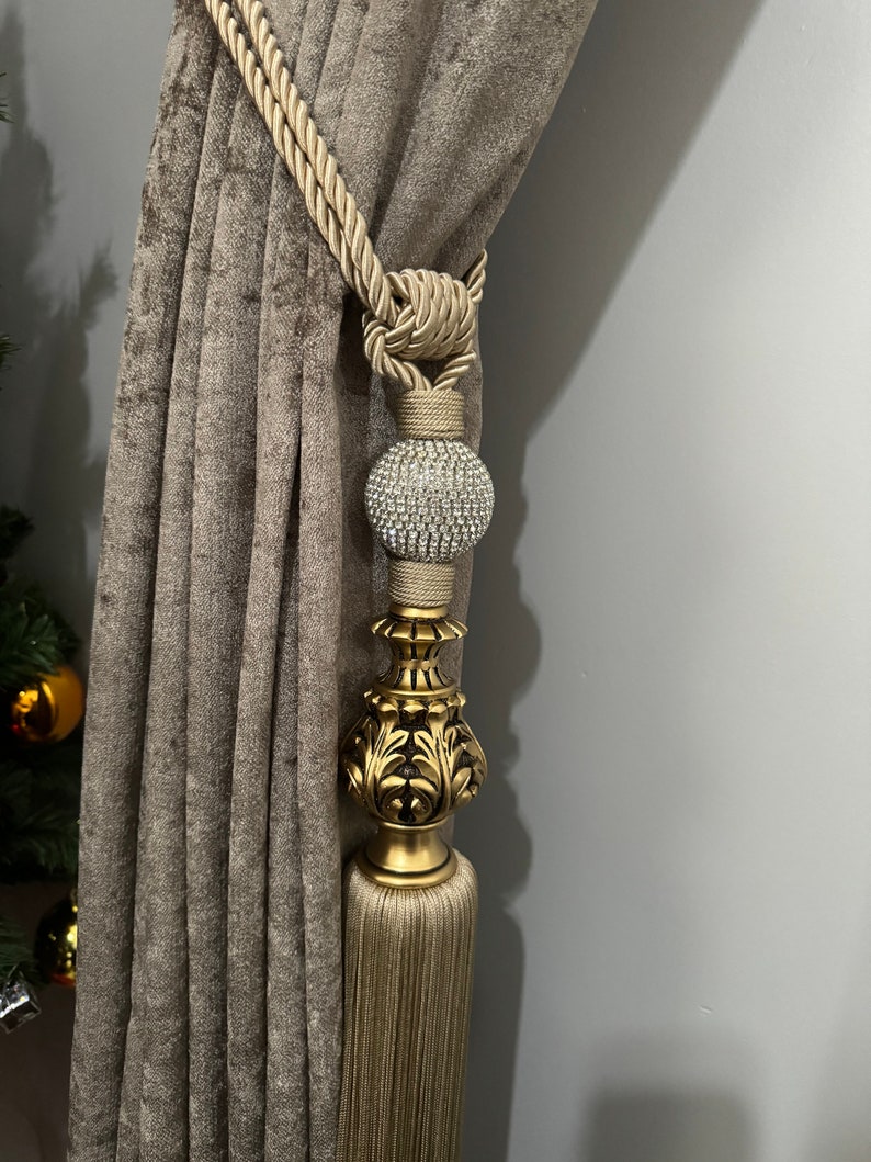May include: A gold curtain tieback with a tassel and a decorative crystal ball. The tieback is made of a thick, braided rope.