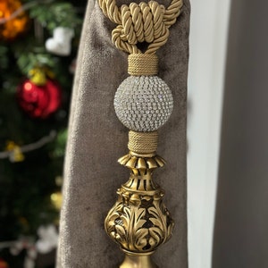 May include: Gold curtain tieback with a decorative tassel and a crystal ball finial. The tieback is made of a braided rope and has a knot at the top.