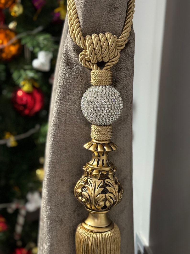 May include: Gold curtain tieback with a decorative tassel and a crystal ball finial. The tieback is made of a braided rope and has a knot at the top.