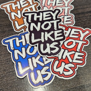 Op de afbeelding: Een stapel stickers met de tekst "They Not Like Us" in vetgedrukte witte letters op verschillende gekleurde achtergronden. De stickers zijn in verschillende kleuren, waaronder rood, blauw, zwart en roze.