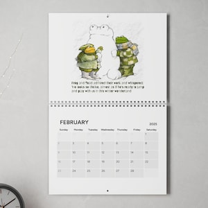 May include: A calendar featuring a February 2025 calendar with a whimsical illustration of two frogs admiring a snowman. The text reads "Frog and Toad admired their work and whispered, 'He looks so lifelike, almost as if he's ready to jump and play with us in this winter wonderland.'"