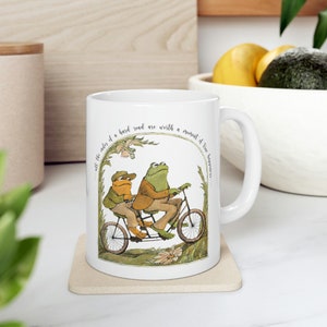 May include: White ceramic mug with a green and brown illustration of two frogs riding a tandem bicycle. The text around the image reads "All the rules of a hard road are worth a moment of true happiness."